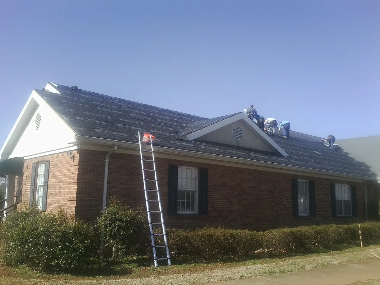 Crew tearing shingles off a brick ranch roof with a ladder set — Metal Roof Repair work by our crew in Bernardston